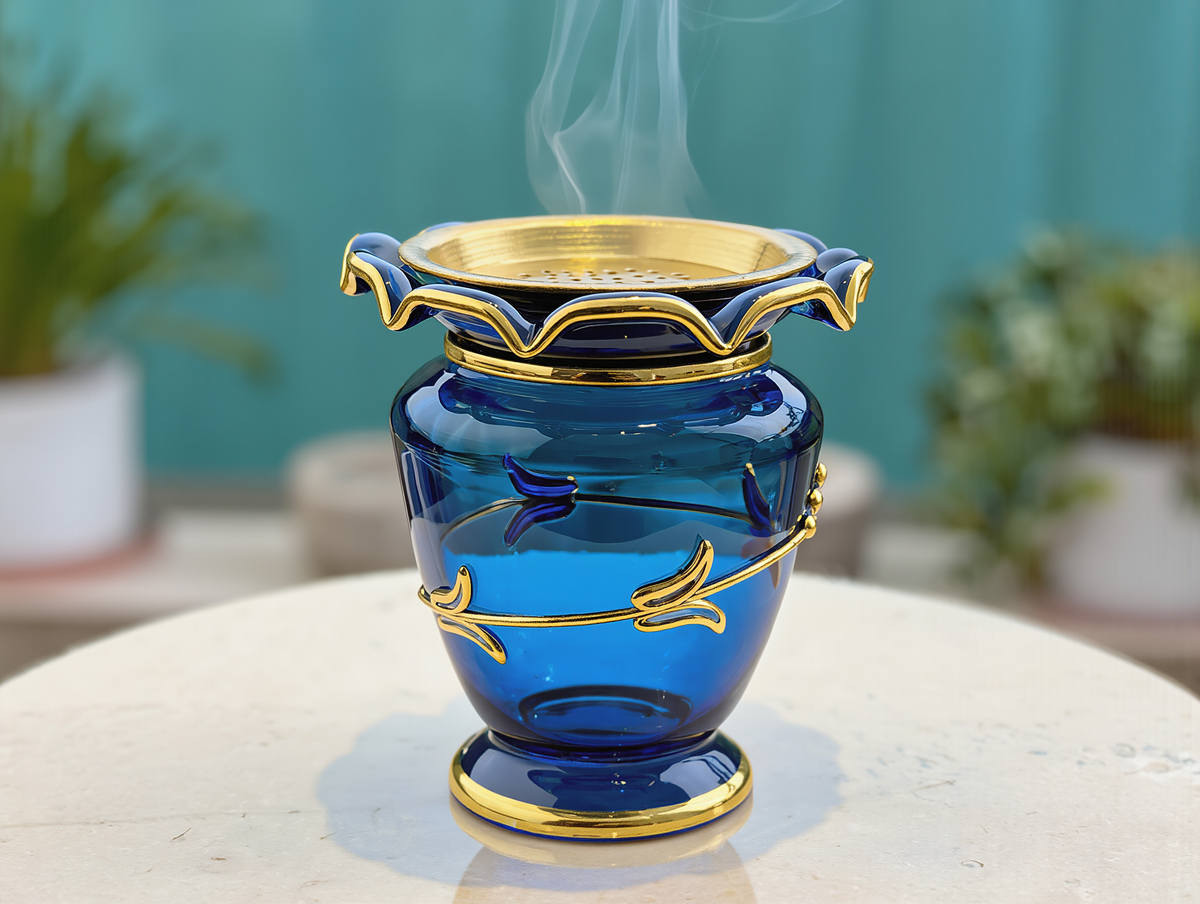 Cobalt blue glass mabkhara with ruffled gold rim and perforated gold tray, gold vine relief, bakhoor smoke rising, on a stone table